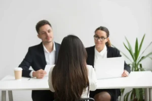 Na imagem uma jovem está de costas sentada de frente para dois entrevistadores durante uma entrevista de emprego.