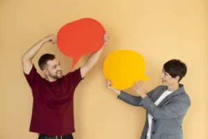 dois jovens sorrindo e segurando papel em formato de balão fazendo alusão a comunicação assertiva