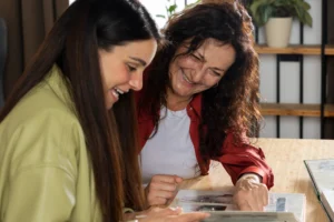 Na imagem, duas mulheres — uma mais jovem e outra mais velha — estão sentadas lado a lado, sorrindo enquanto folheiam um álbum de fotos. O ambiente é acolhedor e iluminado, com plantas e prateleiras ao fundo, transmitindo um clima de aconchego e intimidade. A interação entre elas sugere um momento de lembrança, conexão familiar e carinho ao reviver histórias do passado. Esta imagem é ideal para temas como relações familiares, memórias afetivas, momentos nostálgicos, geração e afeto.