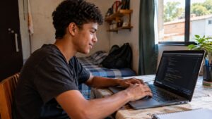 Jovem sentado à mesa de casa, digitando em um notebook com editor de código aberto na tela, aprendendo programação sozinho